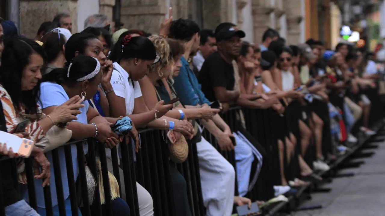 Fila de personas apoyadas en vallas metálicas negras a lo largo de una calle colonial.